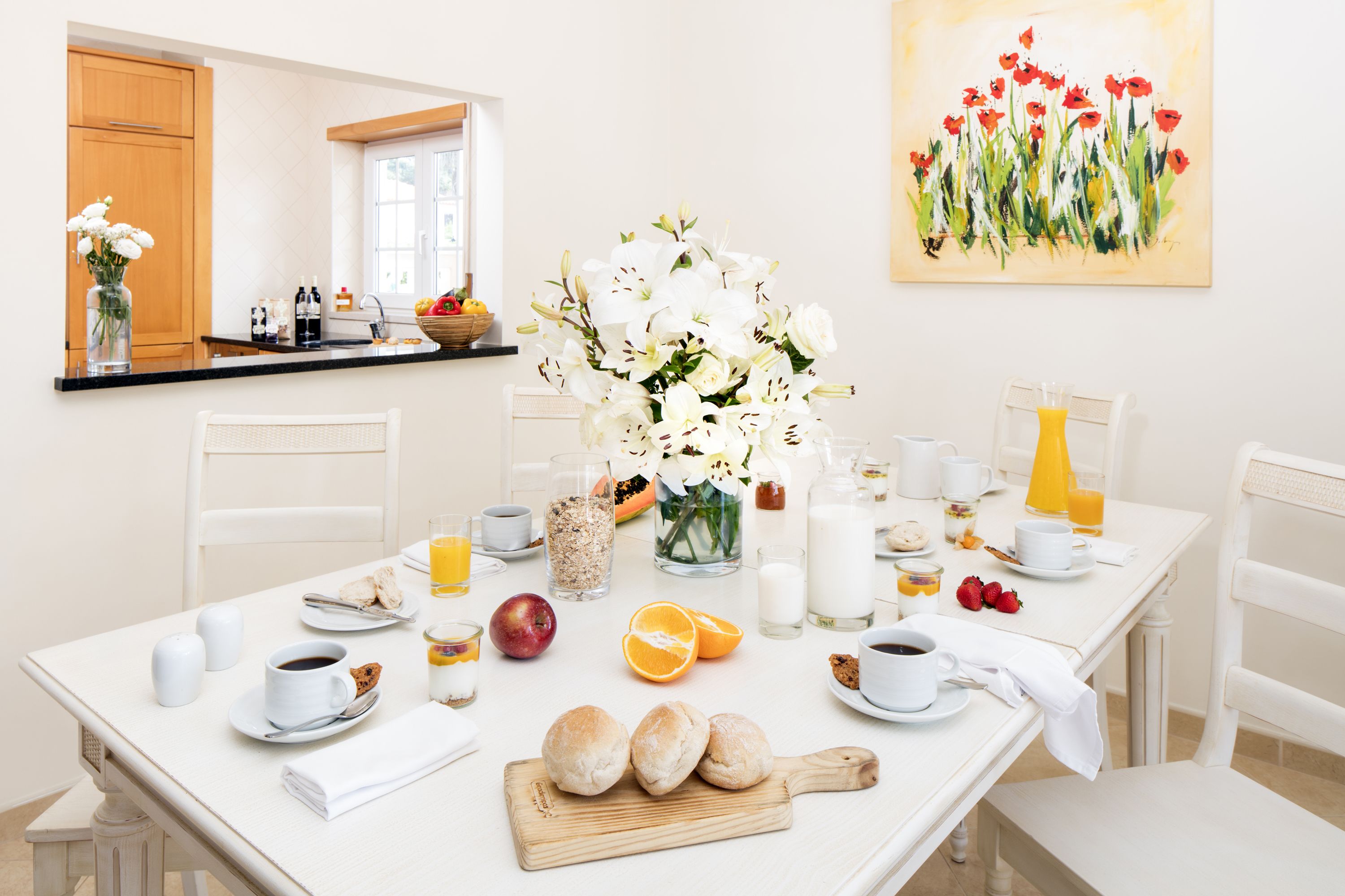 Uma mesa de café da manhã posta com diversos itens, um vaso de flores e um quadro na parede.
