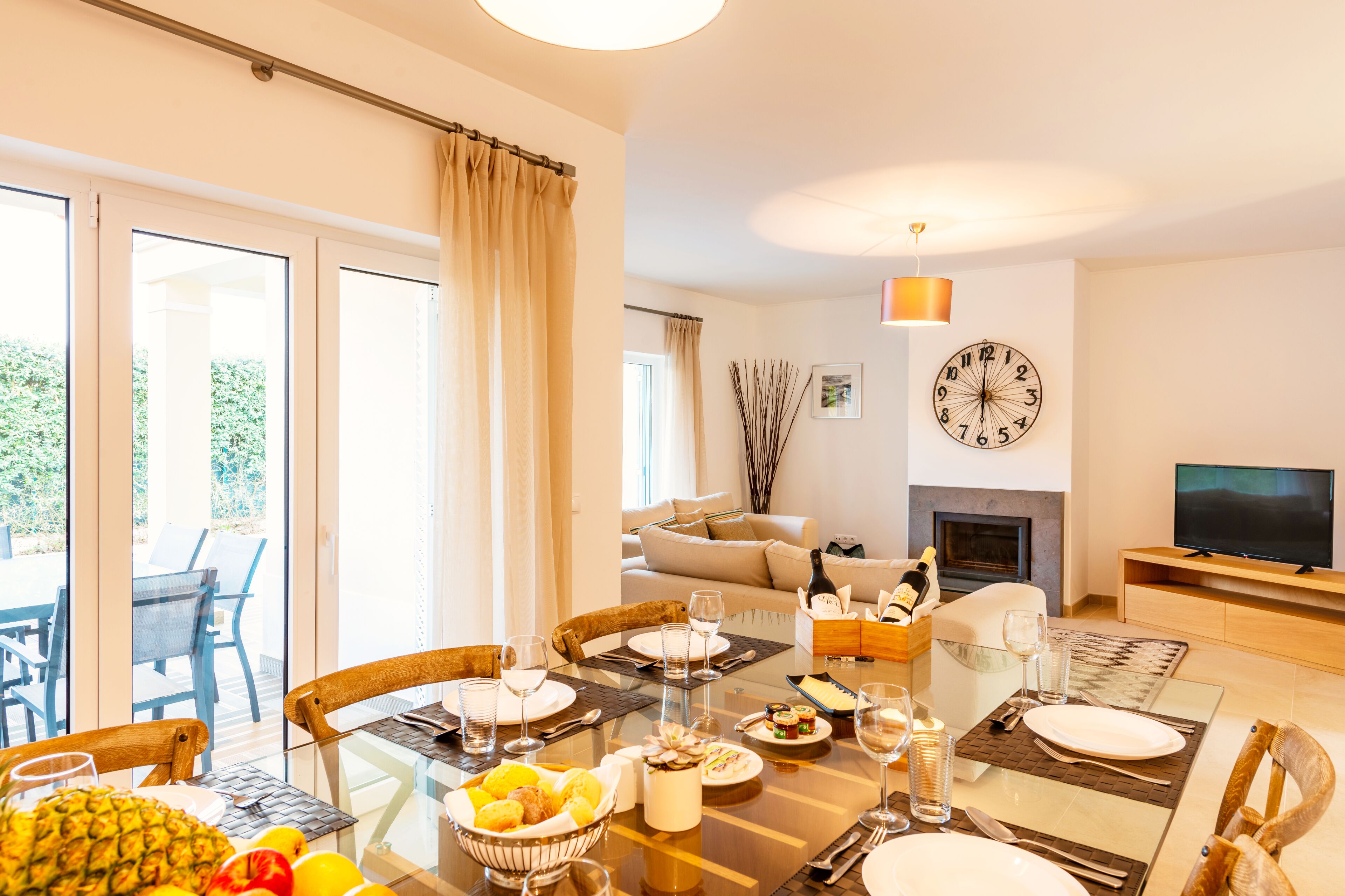 A dining area with a glass table, chairs, and a variety of fruits and dishes. There is a sofa and a TV in the background.