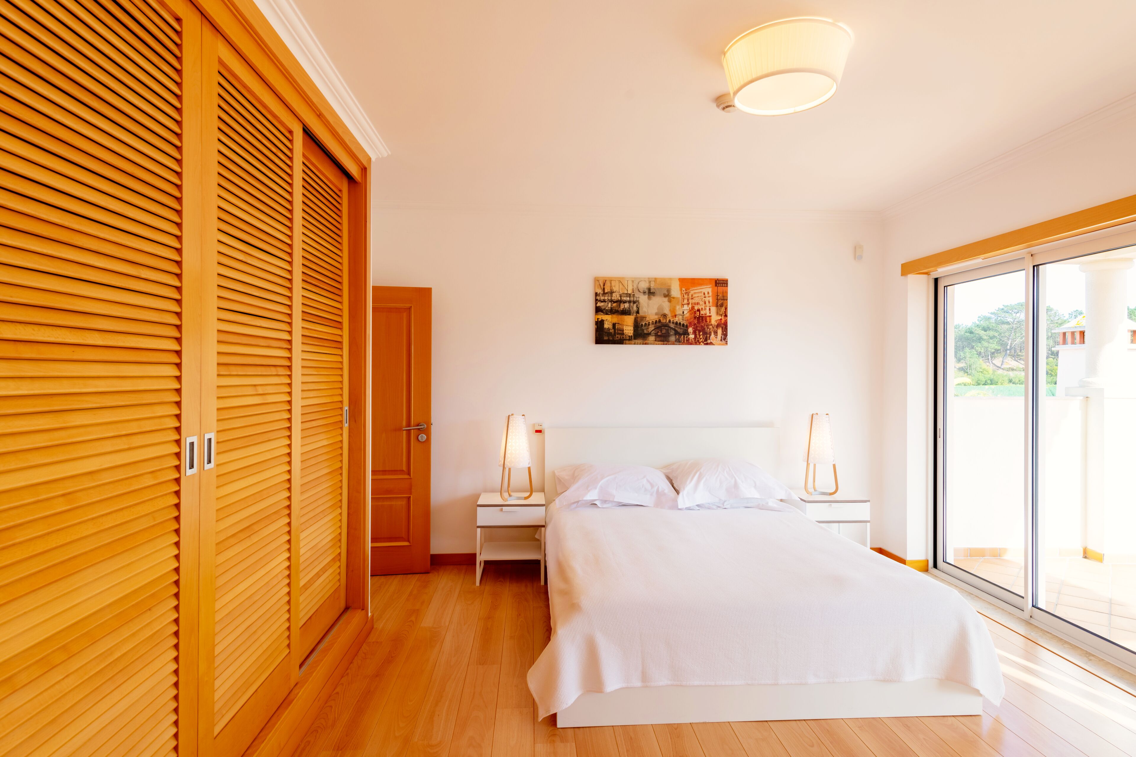 A bedroom with a wooden floor, a bed with white linens, a nightstand with a lamp, and a painting on the wall.