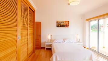 A bedroom with a wooden floor, a bed with white linens, a nightstand with a lamp, and a painting on the wall.