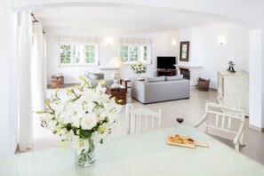 A spacious living room with a sofa, a television, and a fireplace. There is a dining table with a vase of flowers and a plate of food. - The Village Praia d'El Rey Golf & Beach Resort (Obidos)