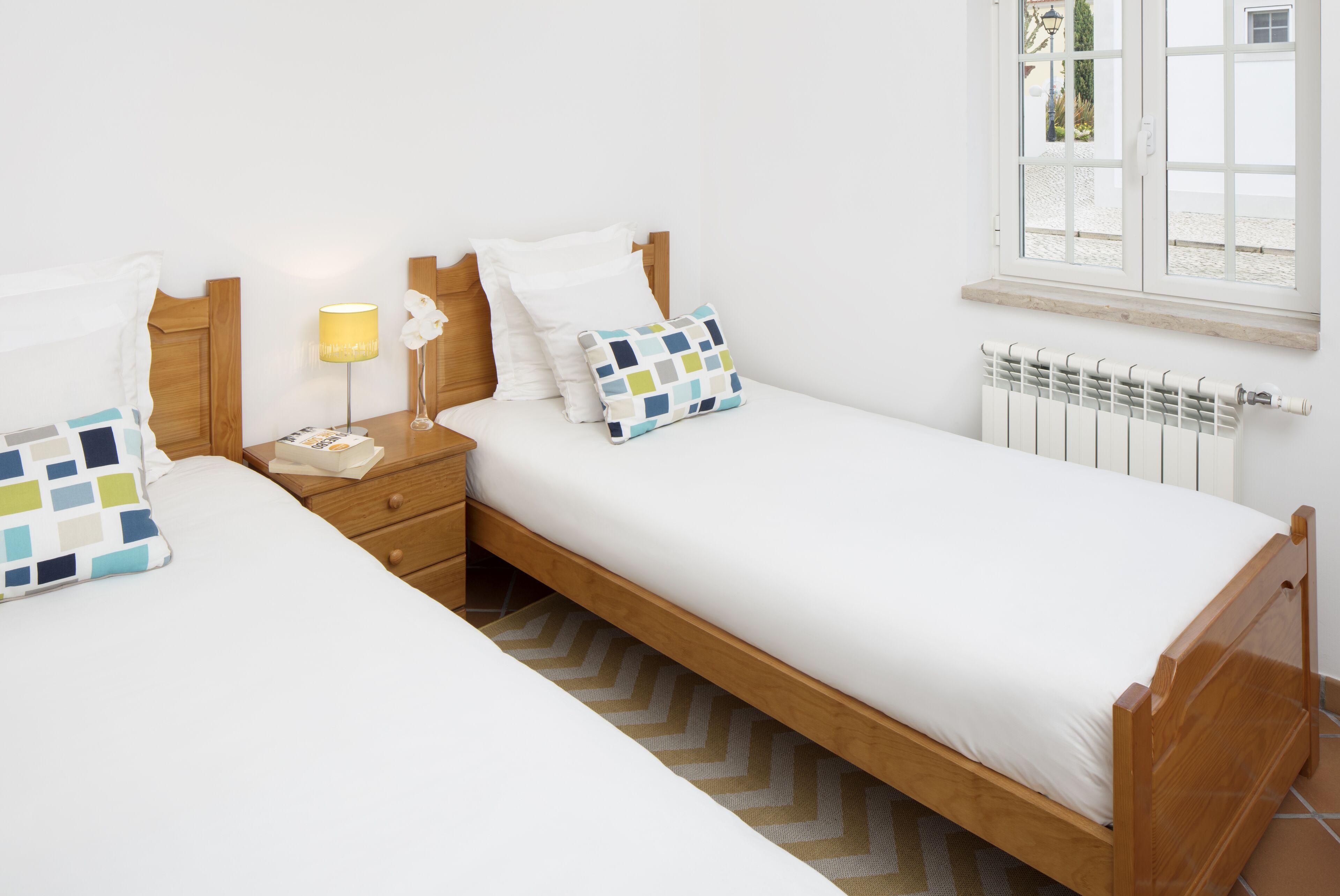 A bedroom with two single beds, wooden headboards, a nightstand with a lamp, and a window with a view of the outside.