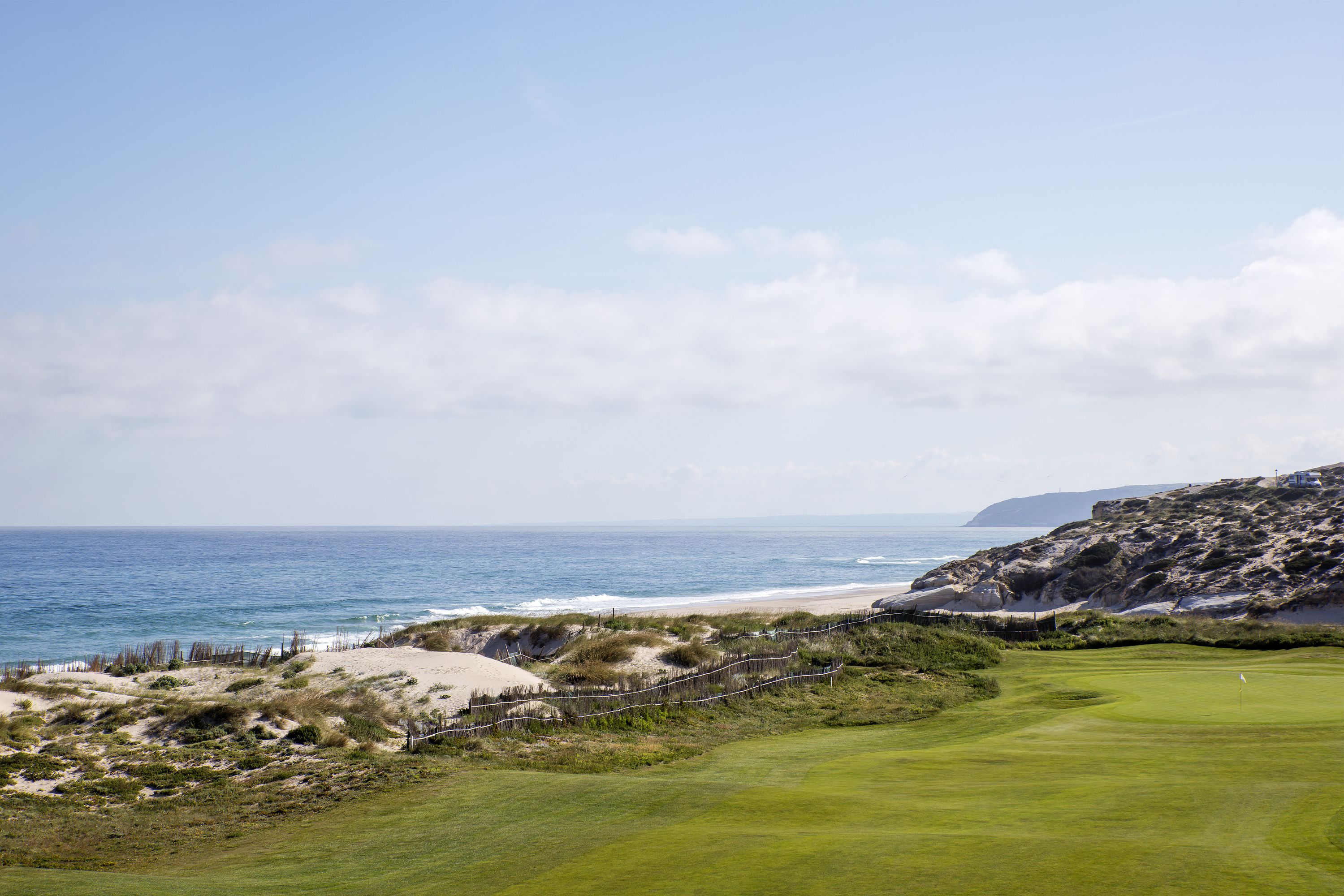 A coastal golf course with a sandy beach, grassy fairways, and a rocky outcrop in the background.
