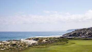 A coastal golf course with a sandy beach, grassy fairways, and a rocky outcrop in the background.