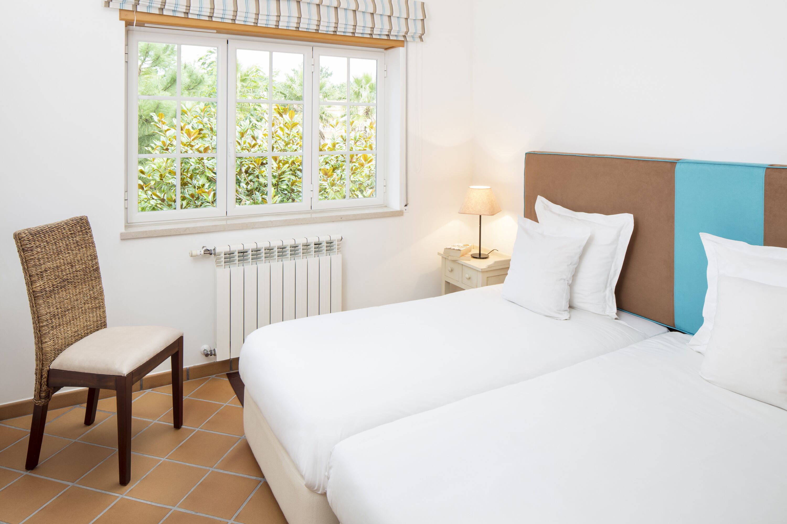 Een hotelkamer met een bed, een stoel, een nachtkastje met een lamp, een raam met uitzicht op groen en een radiator.