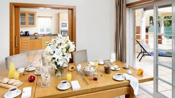 Una mesa de desayuno dispuesta con diversas opciones para el desayuno, un florero con flores y vistas a la cocina y un espacio exterior.