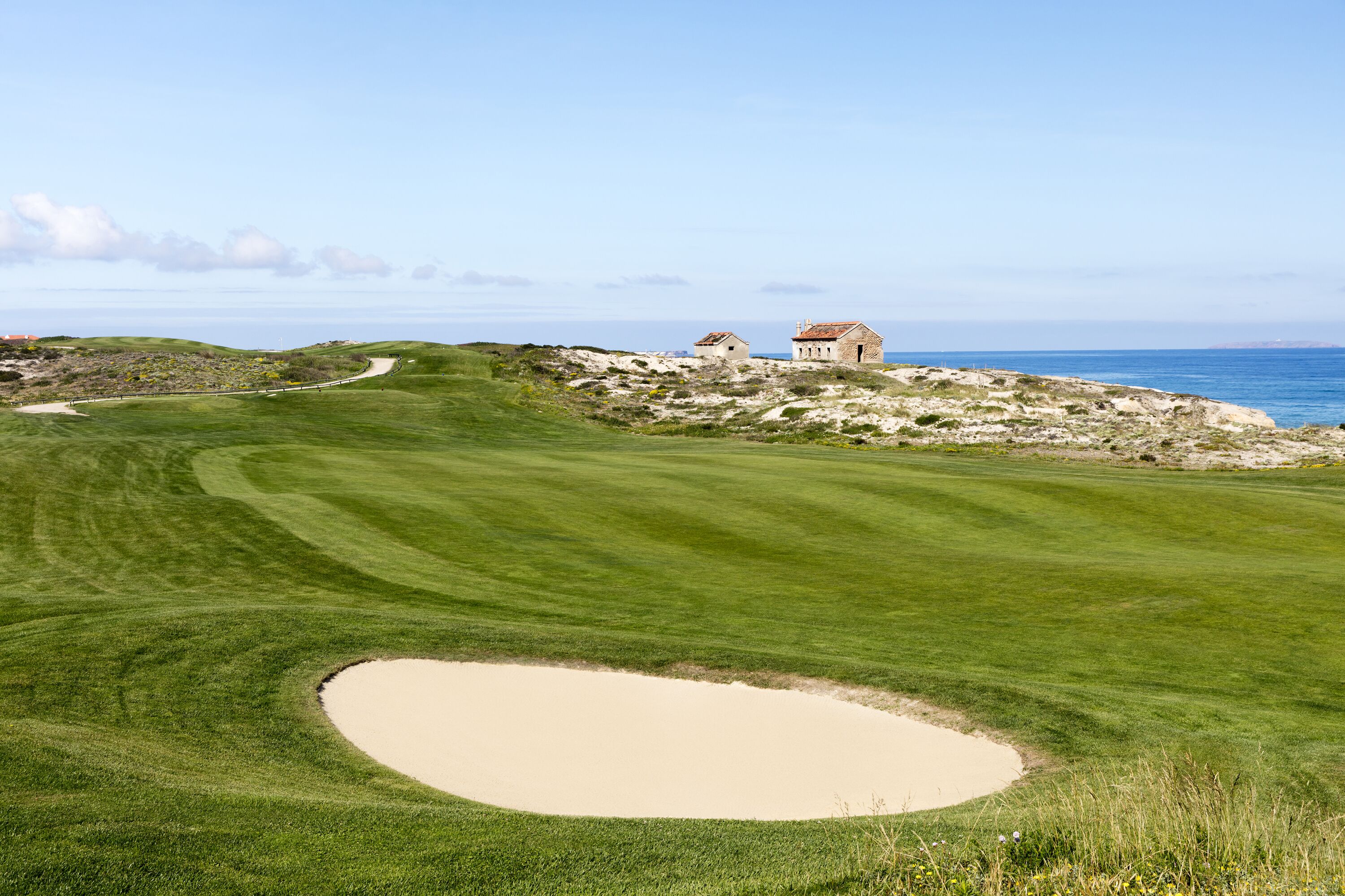 A golf course with a sand bunker, a winding path, and two buildings near the water.