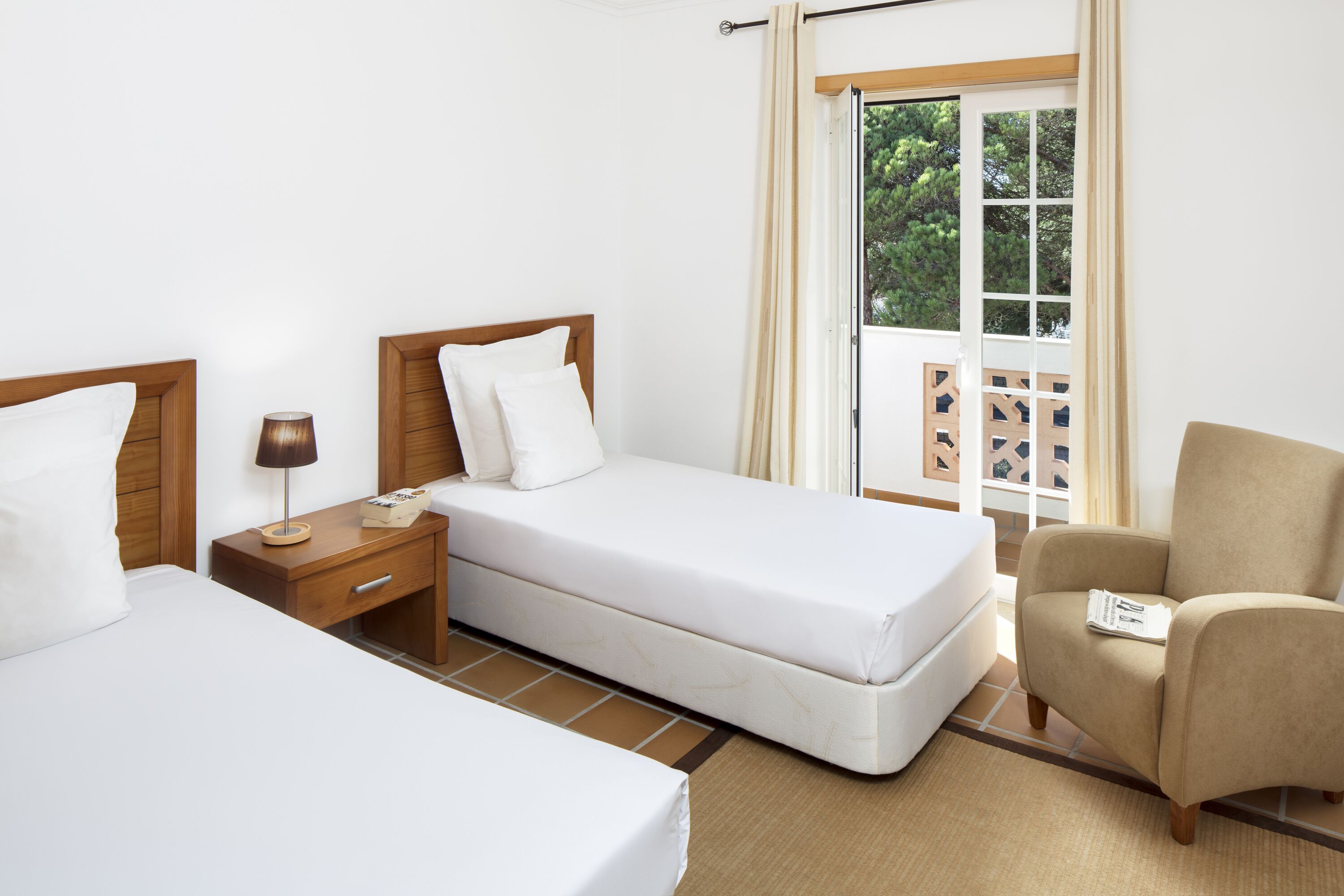 Een hotelkamer met twee bedden, een nachtkastje, een stoel en een raam met uitzicht op groen.