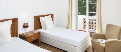 Een hotelkamer met twee bedden, een nachtkastje, een stoel en een raam met uitzicht op groen.