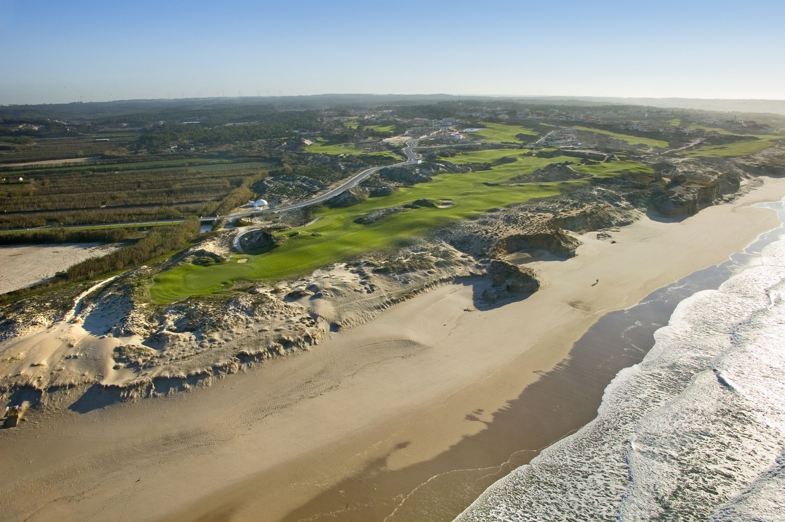 An aerial view of a coastal area with a beach, green fields, and a winding road.