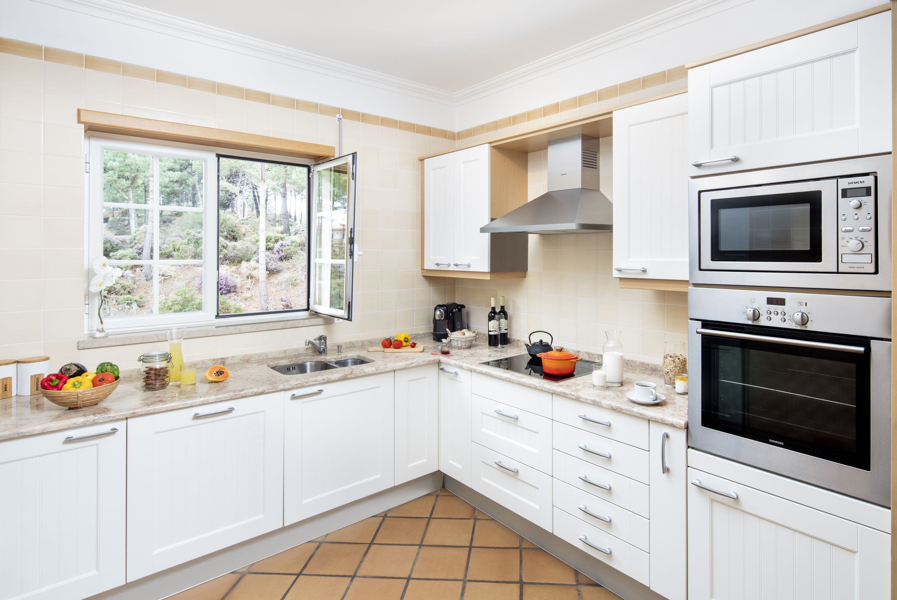 Uma cozinha moderna com armários brancos, forno duplo, micro-ondas e janela com vista para vegetação.