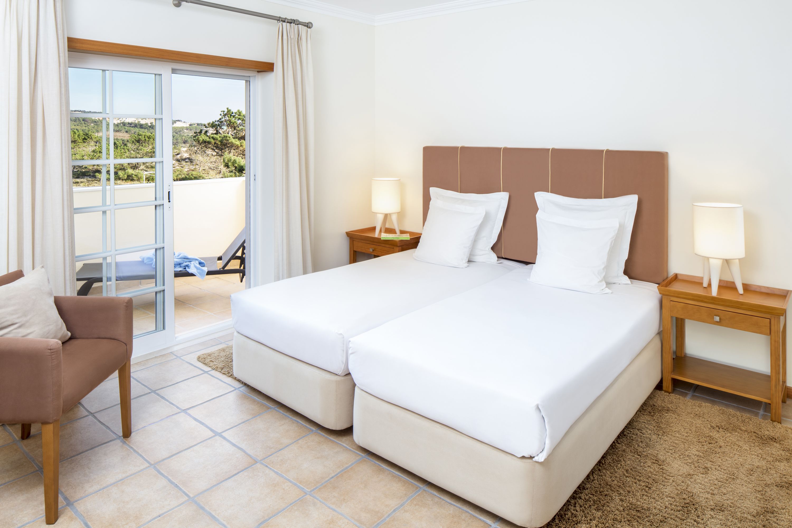 A hotel room with a double bed, two bedside tables with lamps, a chair, and a view of the outdoors through a sliding door.