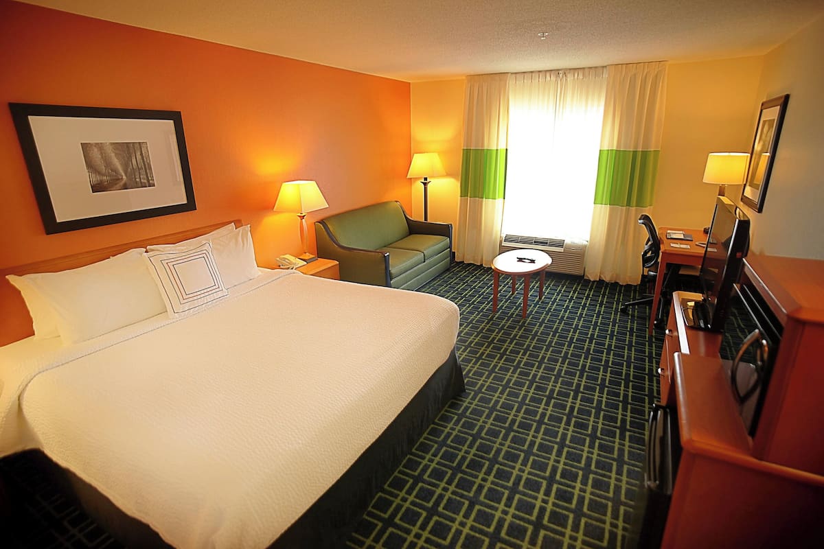 Ein Hotelzimmer mit einem großen Bett, einem grünen Sessel, einem Schreibtisch mit Stuhl und einem kleinen runden Tisch.