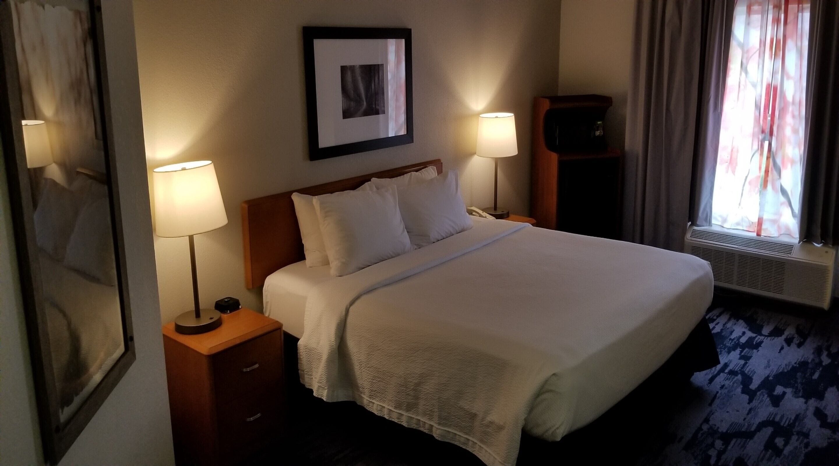 Camera d'albergo con un letto grande, due lampade ai comodini, un quadro appeso al muro e una finestra con tende.