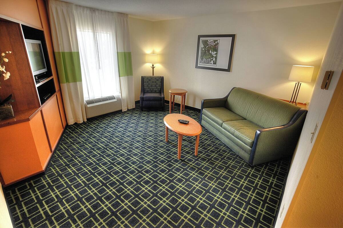 Ein Hotelzimmer mit Teppichmuster, einer Couch, einem Sessel, einem kleinen Tisch, einem Fernseher und einem Fenster mit Vorhängen.