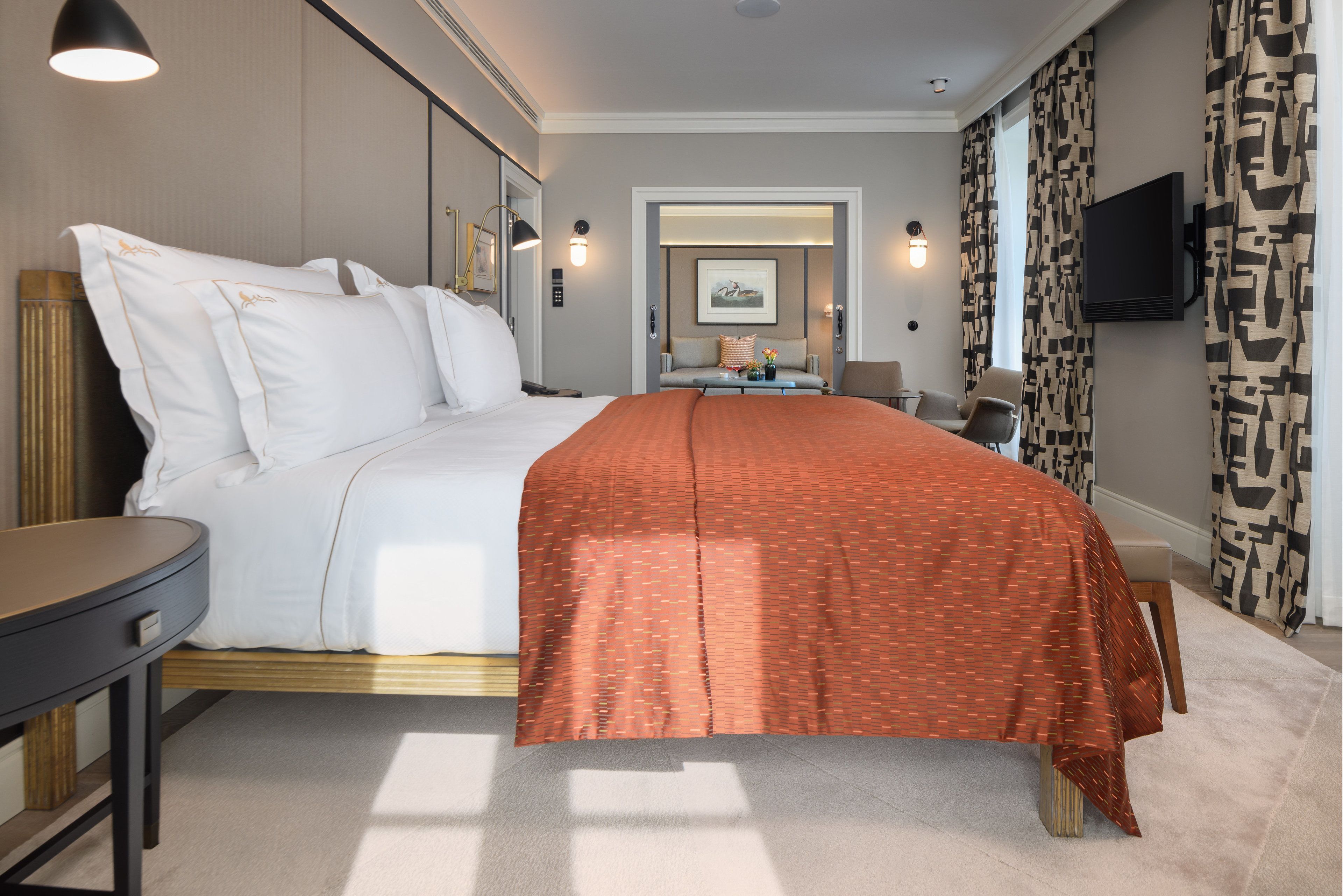 Una habitación de hotel con una cama grande, una mesita de noche, un televisor montado en la pared y un ventanal con cortinas estampadas.
