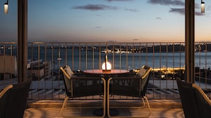 Une terrasse avec une table et des chaises, donnant sur une étendue d’eau, avec les lumières de la ville scintillantes au loin.