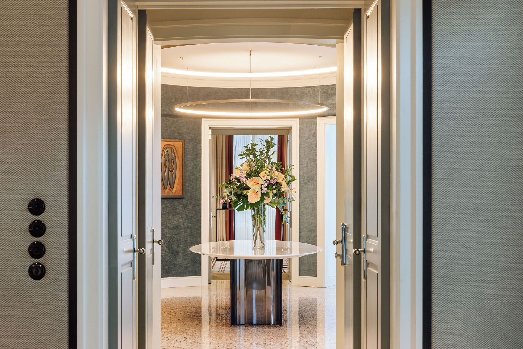 Un pasillo con una mesa redonda y un jarrón de flores, que conduce a una habitación con un ventanal grande y un cuadro en la pared.