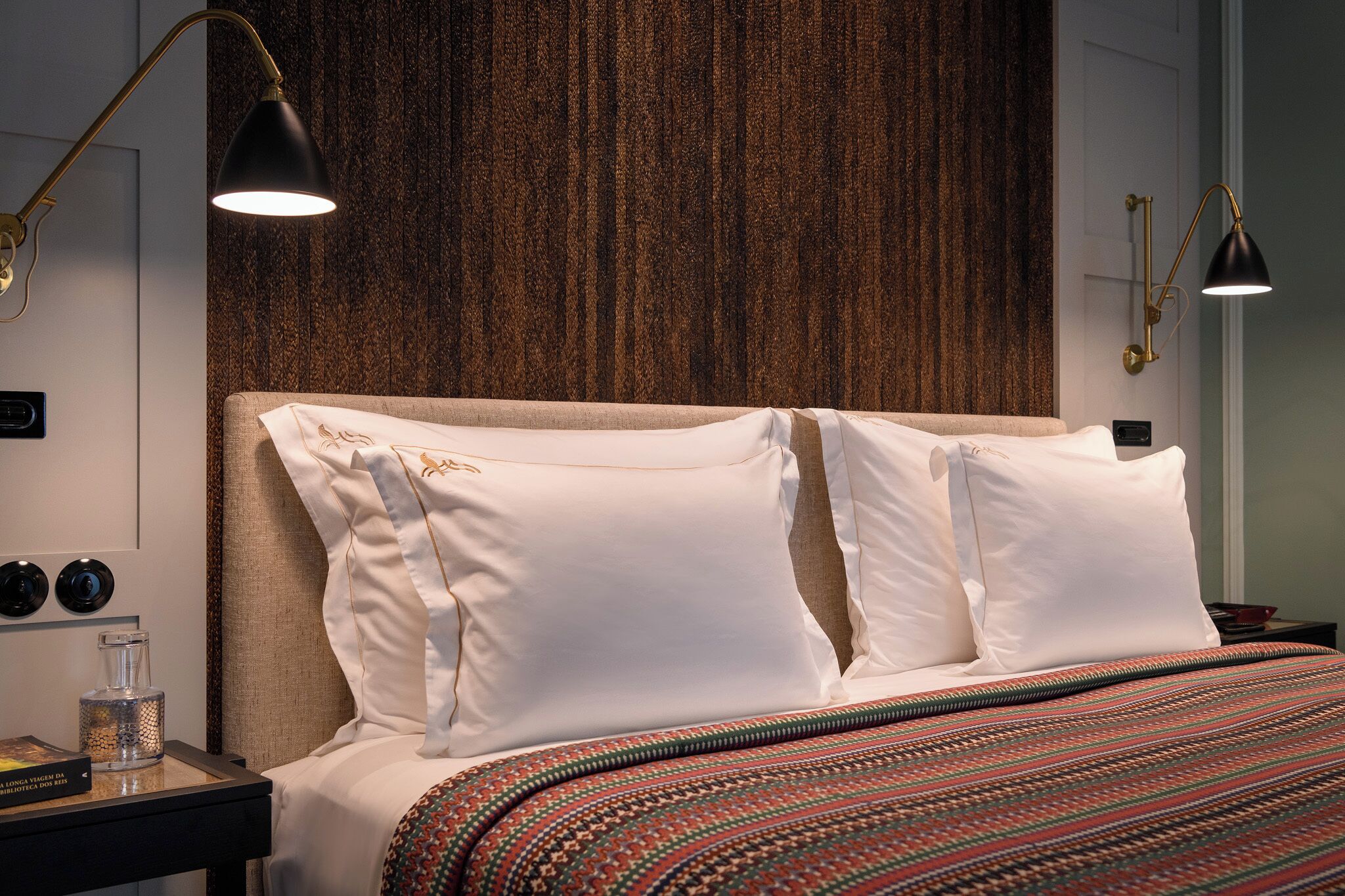 Un lit bien fait, avec du linge de lit blanc et une couvre-lit à motifs, une table de chevet avec une lampe, et un mur orné d’un motif texturé.