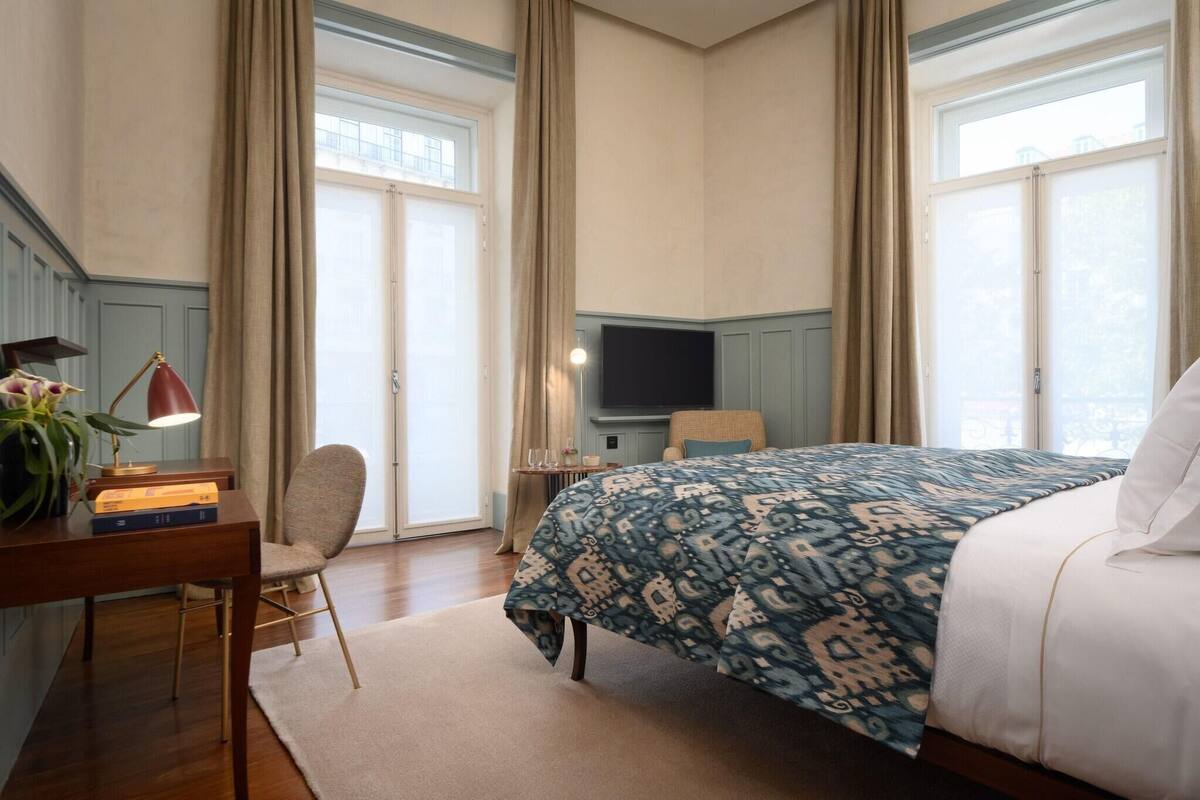 Ett hotellrum med en säng, ett skrivbord med en lampa, en stol, en tv och stora fönster med gardiner.