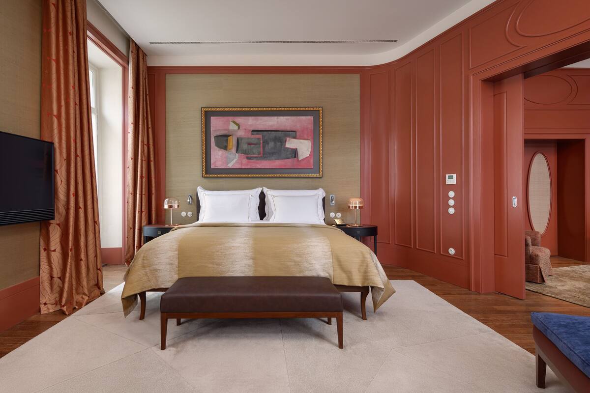 Habitación de hotel con una cama grande, un banco, un televisor, un cuadro en la pared y una puerta con espejo.