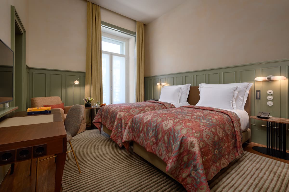 Una habitación de hotel con dos camas, un escritorio, una silla, una mesita de noche y una ventana con cortinas.
