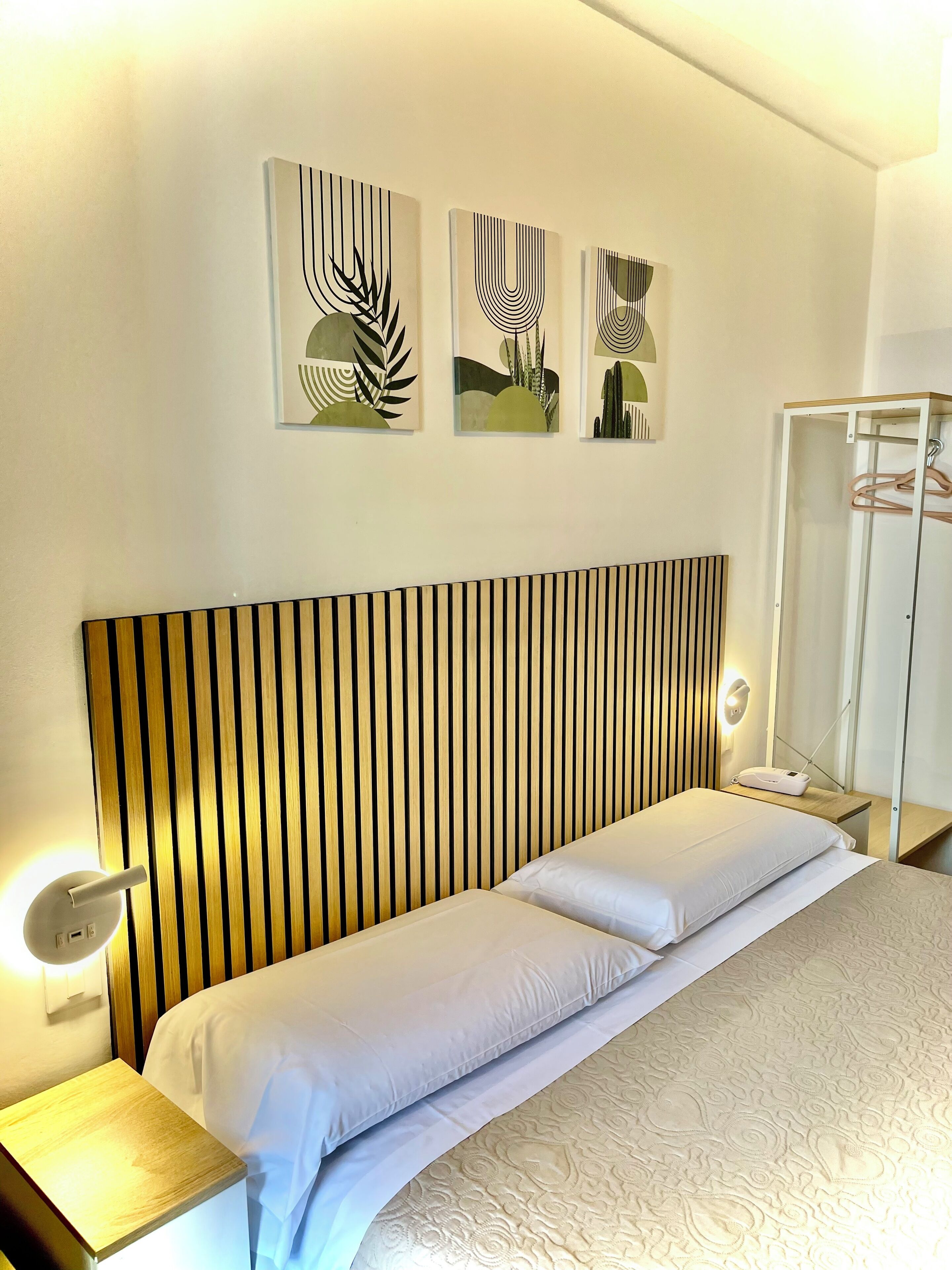 A hotel room with a striped headboard, a bed with white linens, a bedside table with a lamp, and a wardrobe with hangers.
