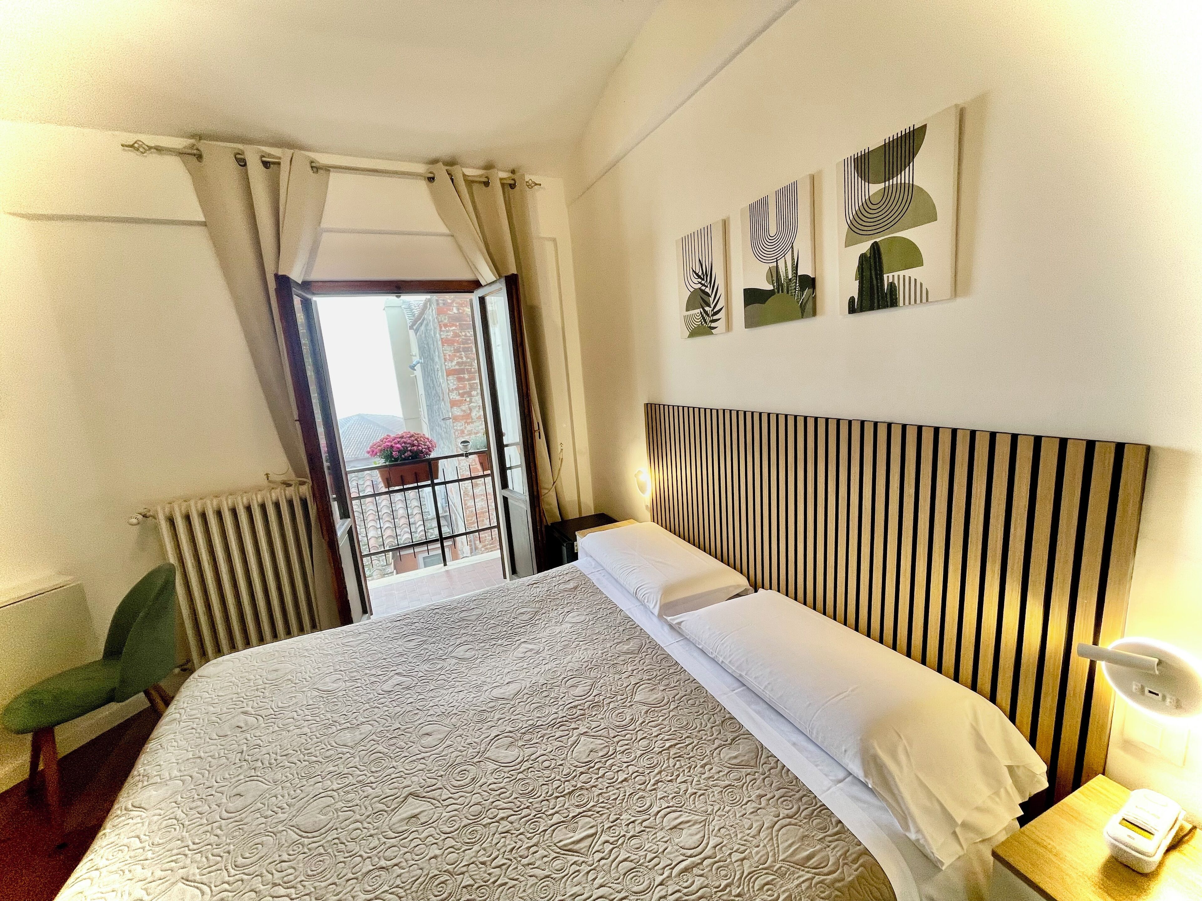 A hotel room with a bed, a chair, a radiator, a window with curtains, and artwork on the wall.