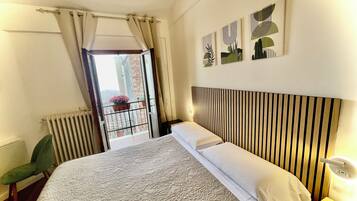 A hotel room with a bed, a chair, a radiator, a window with curtains, and artwork on the wall.