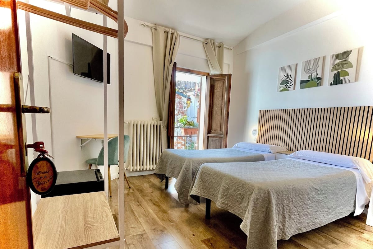 Une chambre d’hôtel avec deux lits, un plancher en bois, un radiateur, un téléviseur fixé au mur et une fenêtre avec des rideaux.