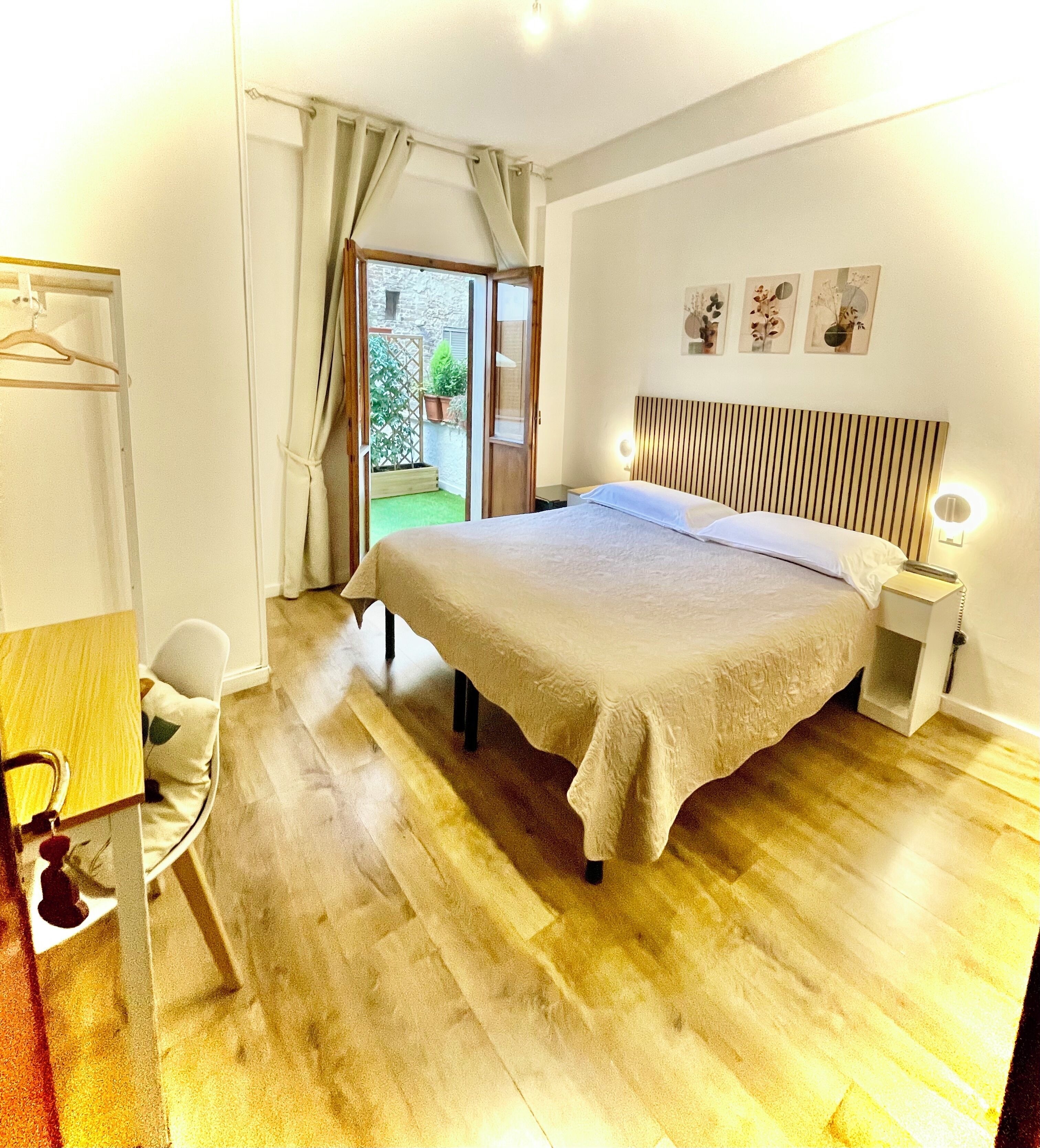A bedroom with a bed, bedside tables, a desk, and a chair. There is a view of a garden through a door.