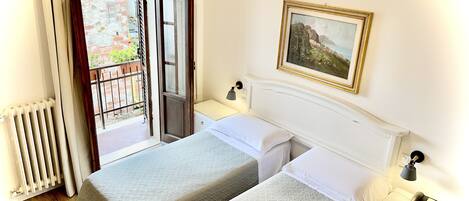 Habitación de hotel con dos camas, un radiador, una mesita de noche con lámpara, un cuadro en la pared y vistas a un balcón con pared de ladrillo y vegetación.