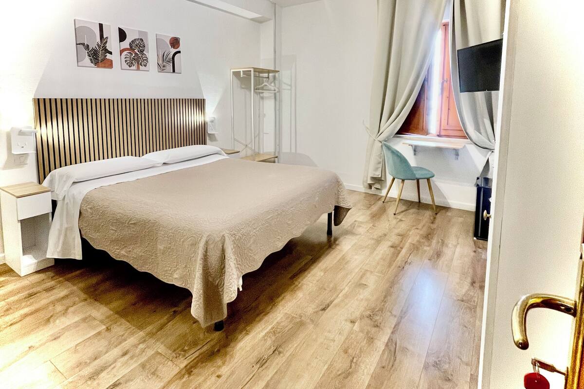 Une chambre moderne avec un plancher en bois, un lit doté d’une tête de lit blanche, une table de chevet, une armoire, un bureau avec une chaise et une fenêtre avec des rideaux.