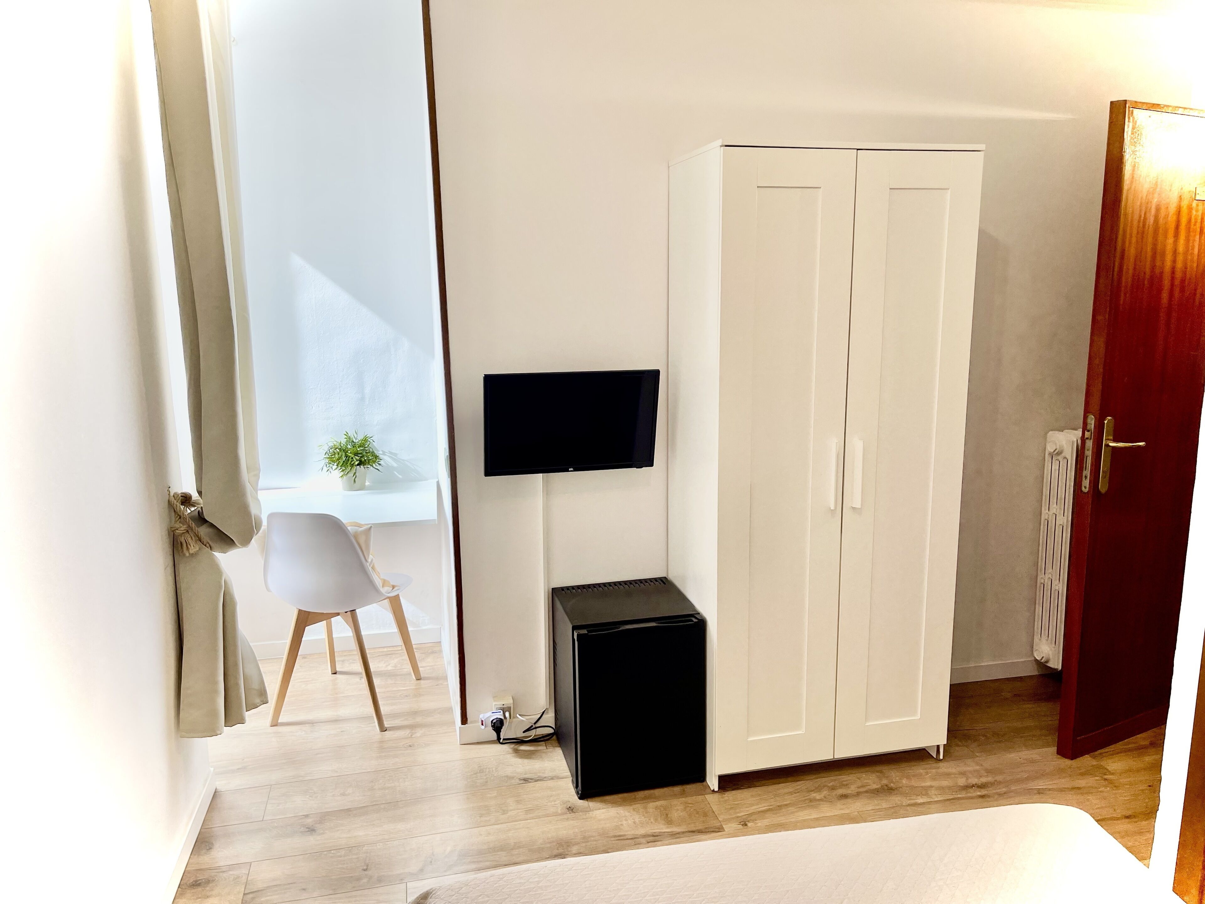 Un dormitorio con una cama, un armario, un escritorio con silla, un pequeño mueble negro y un televisor montado en la pared.