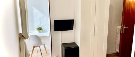 Un dormitorio con una cama, un armario, un escritorio con silla, un pequeño mueble negro y un televisor montado en la pared.