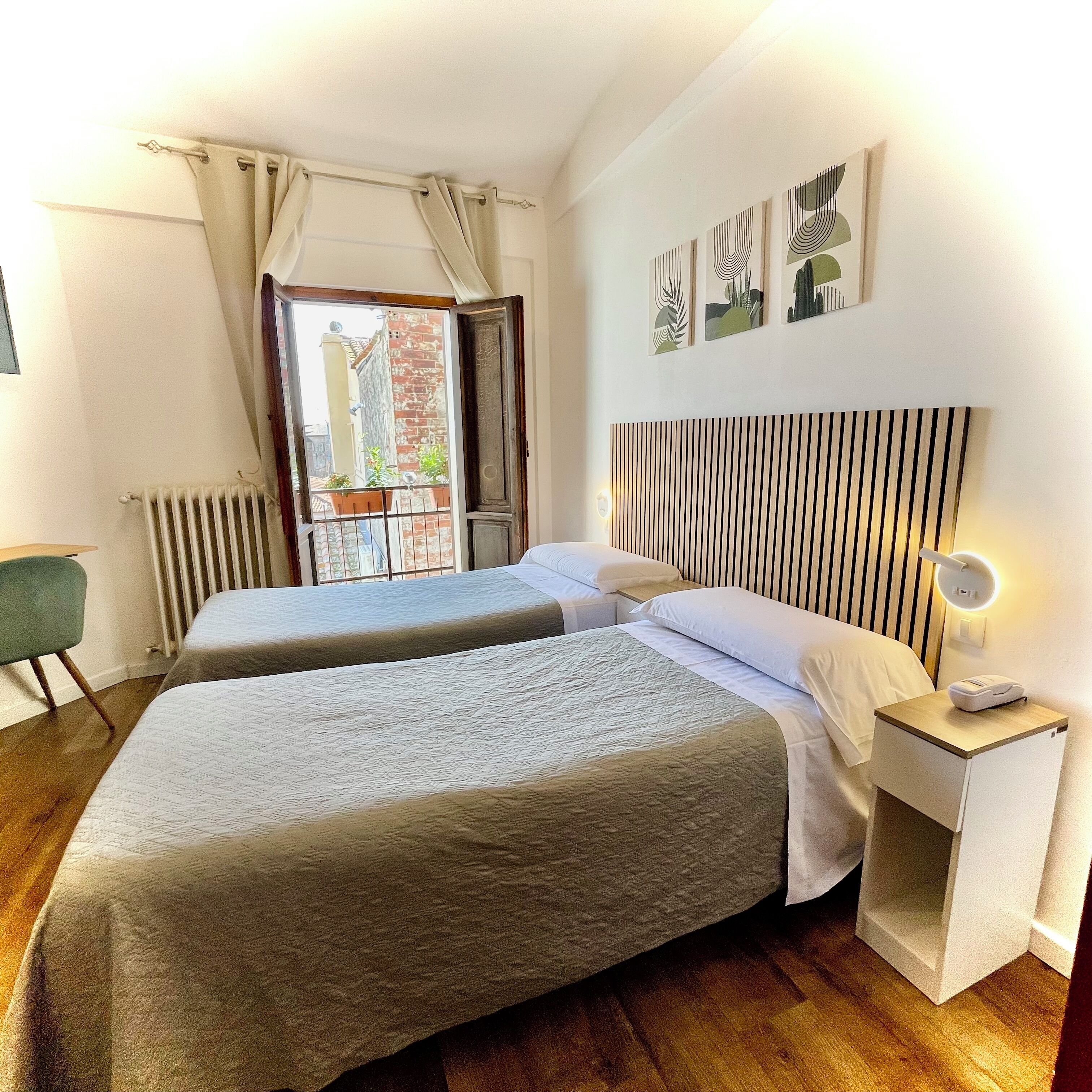 Habitación de hotel con dos camas, cabecera de madera, mesita de noche con lámpara y vista al balcón exterior.