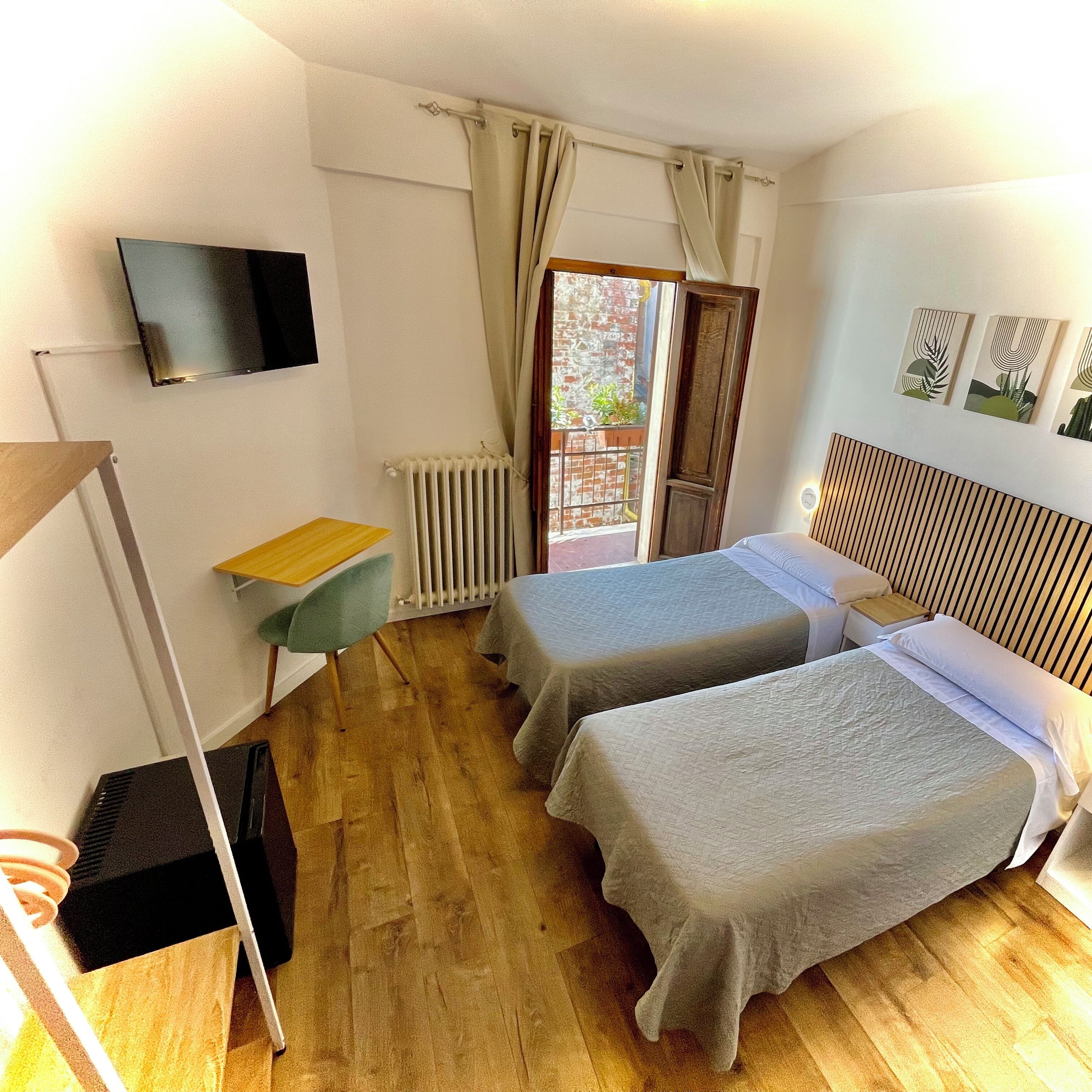 A room with two beds, a desk, a radiator, a TV mounted on the wall, and a window with curtains.