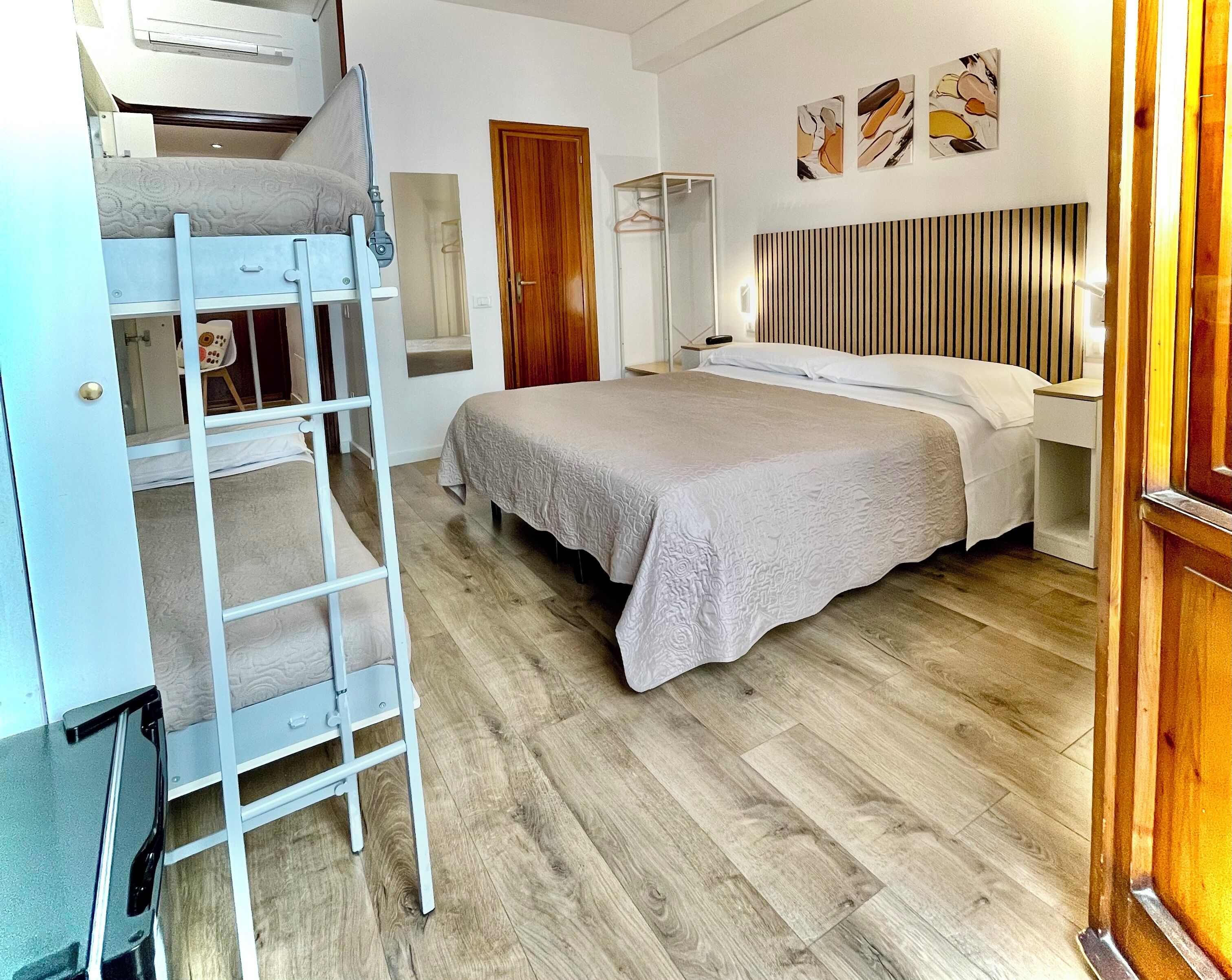 A bedroom with a bunk bed, a bed with a headboard, wooden flooring, a wardrobe, and a mirror.