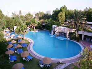 Une grande piscine incurvée, dotée de chaises longues et de parasols, entourée de verdure et agrémentée d’une cascade.