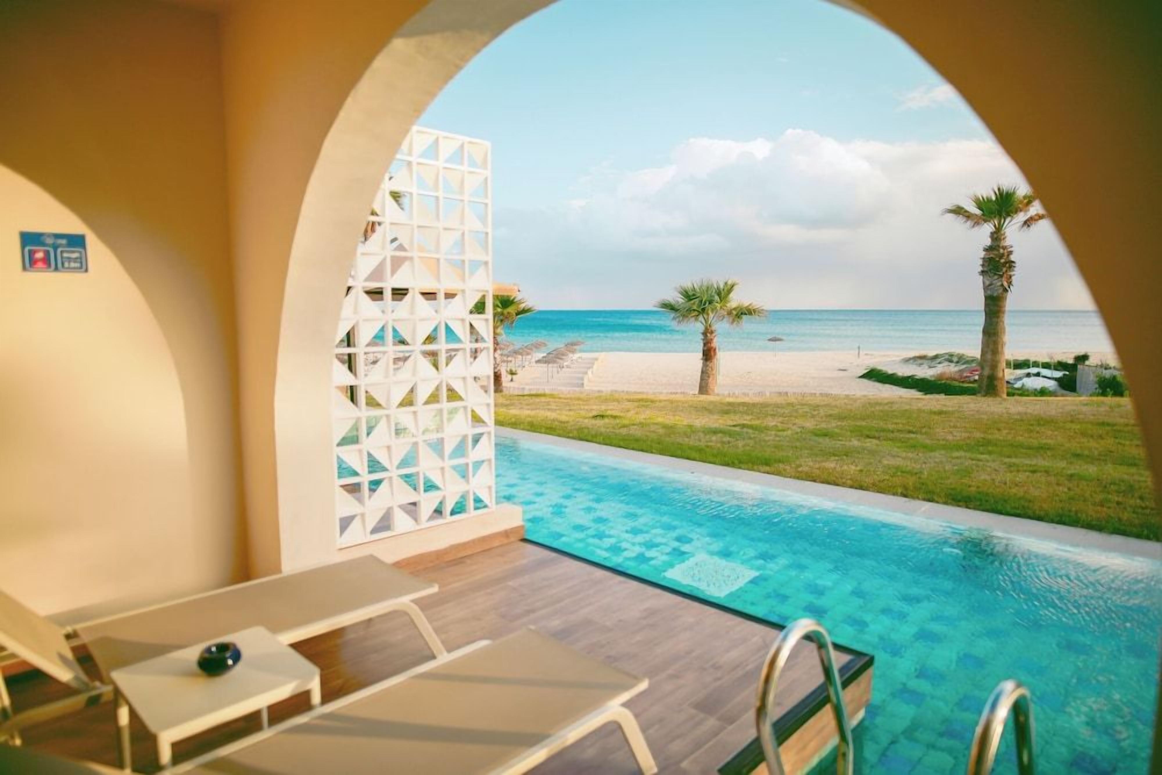 A poolside area with a view of the beach, a palm tree, and a white lattice structure.