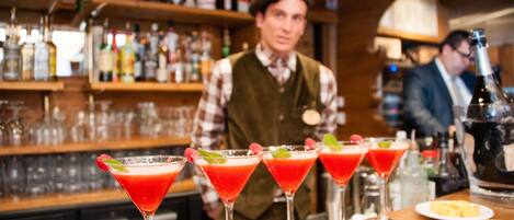 A bar with multiple red cocktails garnished with mint and strawberries, a bartender in uniform, and shelves stocked with various bottles.