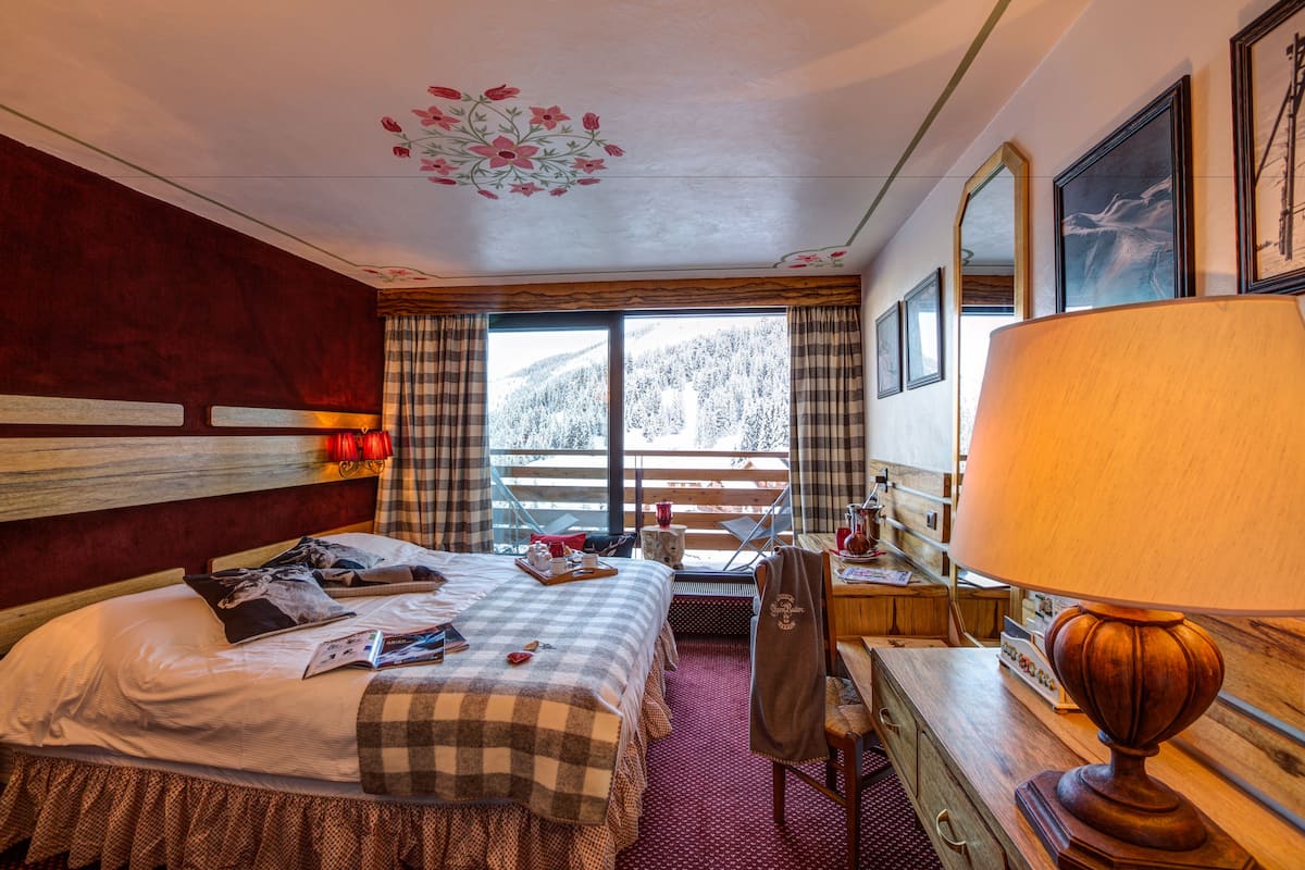 Une chambre d’hôtel comprenant un lit, un bureau, une chaise, une lampe, une fenêtre donnant sur les montagnes et un plafond décoré de motifs floraux.