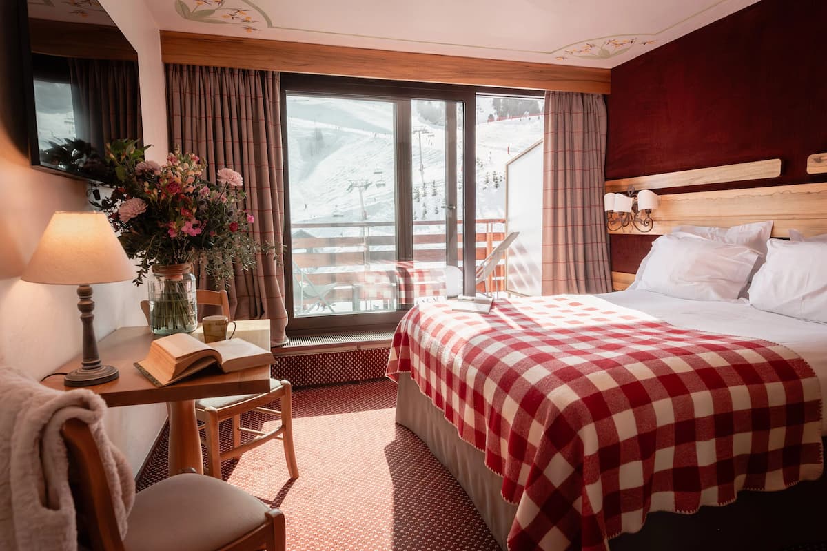 Une chambre d’hôtel avec un lit, un bureau avec une lampe, une chaise, une fenêtre donnant sur une piste de ski et une couvre-lit à carreaux rouges et blancs.