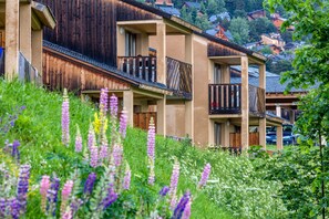 A row of wooden and beige buildings with balconies, surrounded by lush greenery and wildflowers.