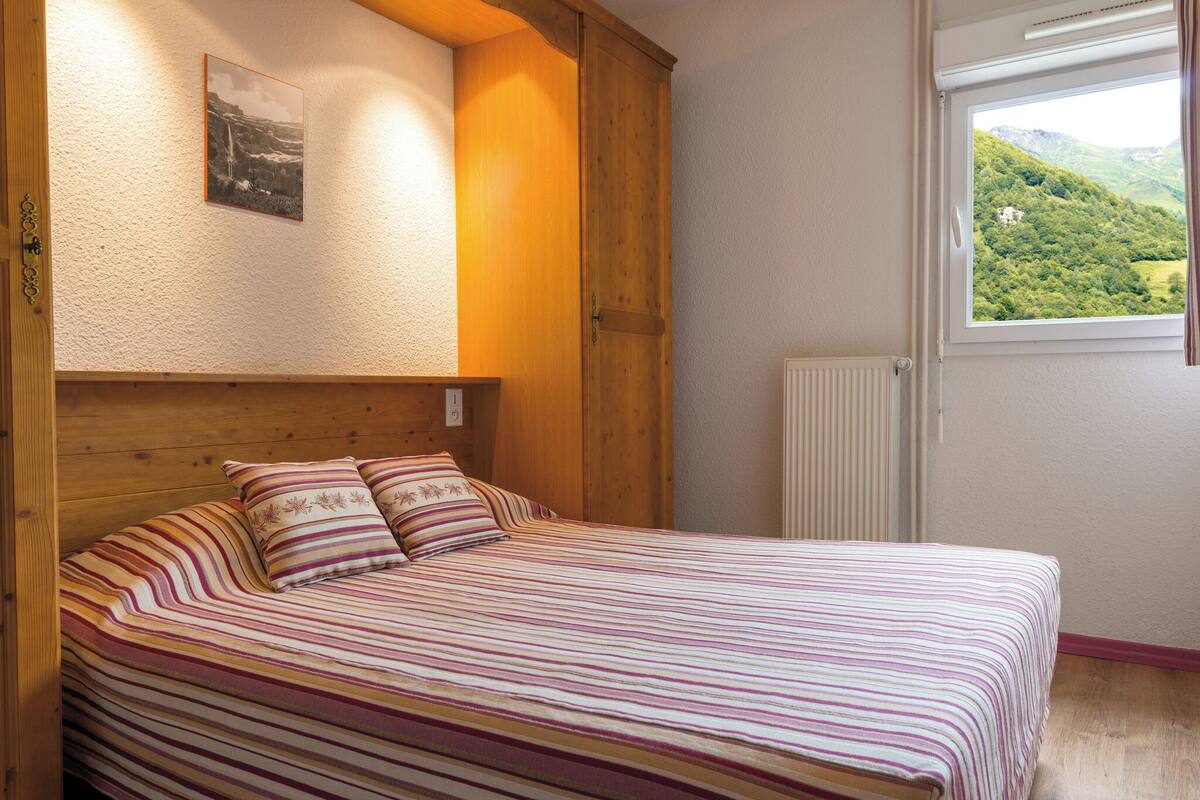Una cama con ropa de cama a rayas, un cabecero de madera, un cuadro enmarcado en la pared y una ventana con vistas a la vegetación.