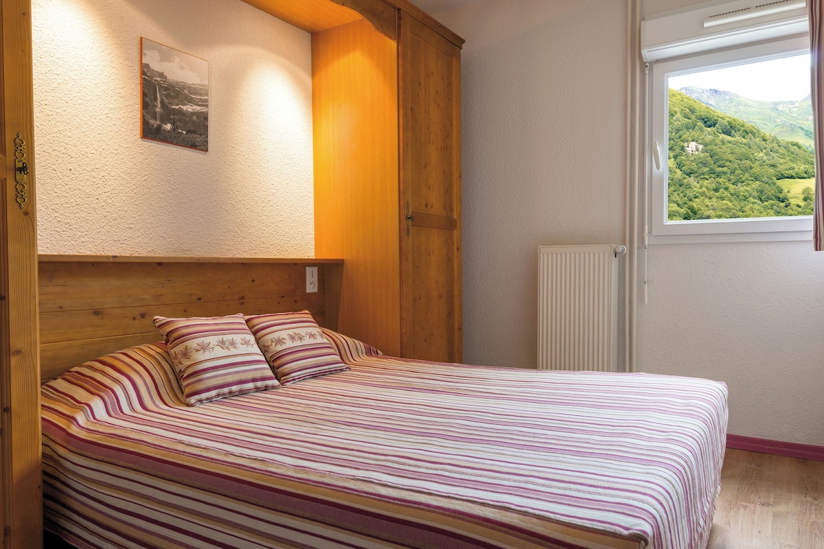 Una cama con ropa de cama a rayas, un cabecero de madera, un cuadro enmarcado en la pared y una ventana con vistas a la vegetación.
