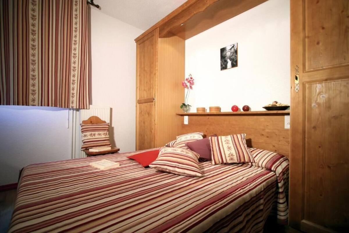 Una cama con ropa de cama a rayas, cabecera de madera, una pequeña estantería con objetos decorativos y una ventana con cortinas.