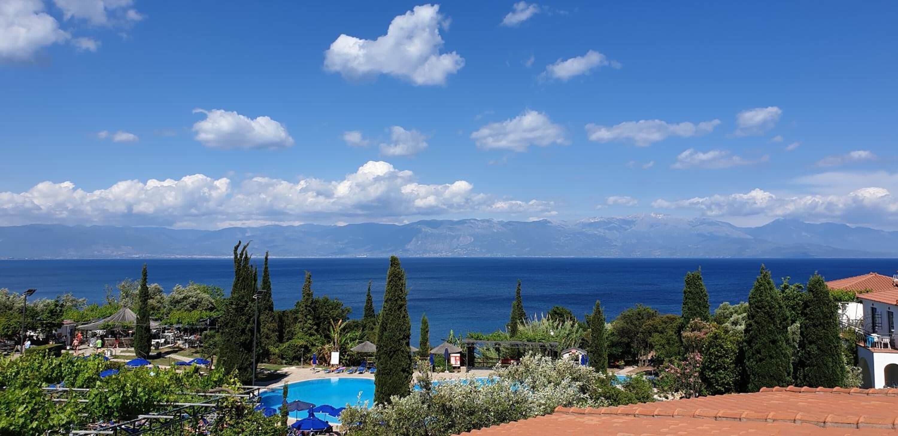 A coastal resort with a swimming pool, beach umbrellas, and a view of the sea and mountains.
