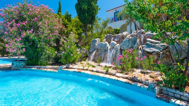 Una piscina con agua azul cristalina, rodeada de vegetación exuberante y una cascada artificial de roca.