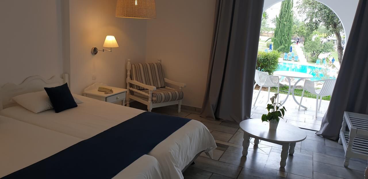 A hotel room with a bed, a nightstand, a chair, a small table with a plant, and a view of an outdoor pool area.